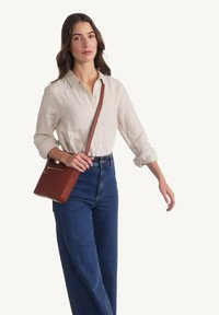 Sac en bandoulière en cuir brun avec une poche frontale zippée, texture lisse, forme rectangulaire. Modèle en chemise claire et jean large.