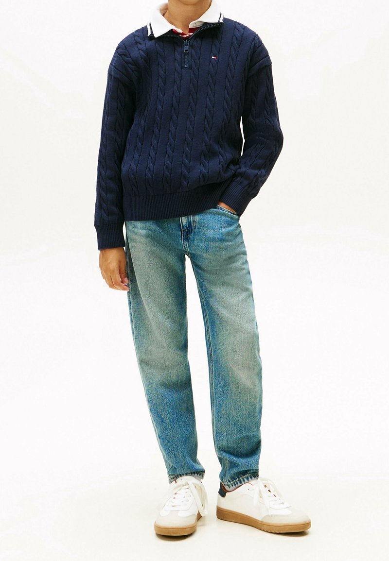 Enfant portant un pull en maille torsadée bleu marine, une chemise blanche à col, un jean bleu clair et des baskets blanches avec semelles en gomme, debout avec une main dans la poche.