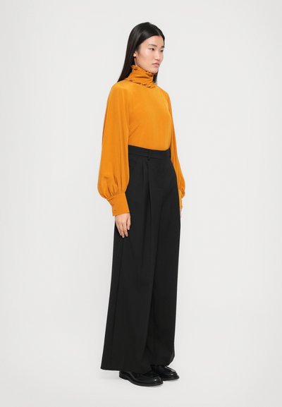 Blusa arancione a collo alto con colletto arricciato e maniche lunghe; abbinata a pantaloni neri a gamba larga e scarpe nere.