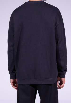 Persoon in een eenvoudige donkerblauwe lange mouwen sweatshirt en bijpassende donkere broek, gezien vanaf de achterkant tegen een lichte achtergrond.