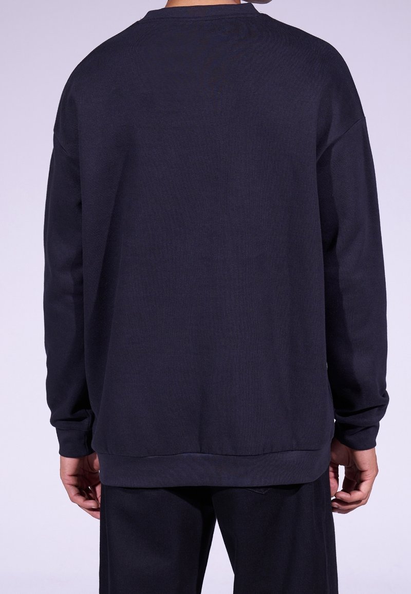 Eine Person trägt einen schlichten, dunkelblauen Langarm-Pullover und passende dunkle Hosen, von hinten vor einem hellen Hintergrund abgebildet.