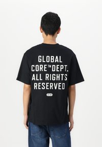 Svart t-skjorte med fet hvit tekst på ryggen: "GLOBAL CORE™ DEPT. ALL RIGHTS RESERVED" med "EST.90" under. Korte ermer, avslappet passform.