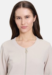 Veste zippée beige clair, tissu texturé, col rond, quincaillerie dorée et design minimaliste. Convient pour diverses occasions décontractées.