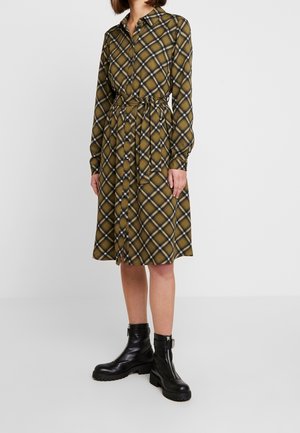 Femme portant une robe-chemise marron à carreaux, manches longues, longueur genou, avec une ceinture nouée à la taille et des bottines noires à fermeture éclair.