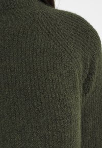 Närbild av en person som bär en mörkgrön texturerad stickad tröja, med fokus på axeln och övre delen av ryggen.