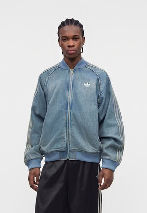 Ung mand med flettet hår iført en blå denim Adidas lynlåstrøje og sorte Adidas bukser med hvide striber.