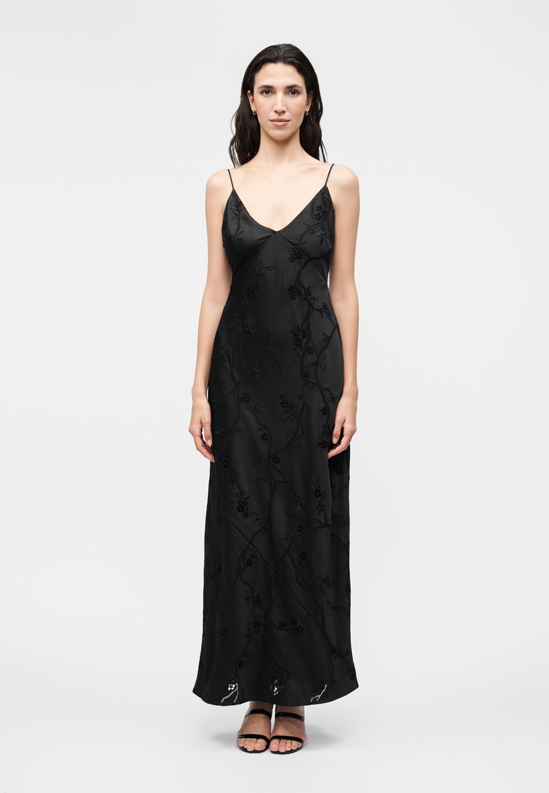 Γυναίκα με μακριά σκούρα μαλλιά που φοράει μαύρο φορεματόμαξι με λεπτές ραντάκια και κεντήματα με λουλούδια και μαύρα σανδάλια με λουράκια, στέκεται μπροστά σε λευκό φόντο.