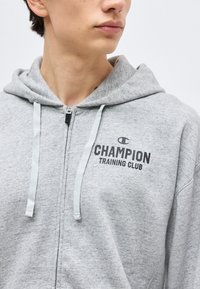Persoană tânără purtând un hanorac cu fermoar de culoare gri deschis, cu logo-ul "Champion Training Club" pe piept, orientată ușor spre dreapta, decupată la nivelul gâtului și umerilor.