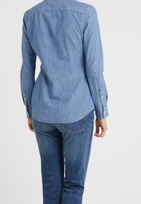 Camisa de mezclilla en azul claro, diseño de botonadura, mangas largas con puños abotonados, dobladillo ligeramente curvado, combinada con jeans de mezclilla oscura.