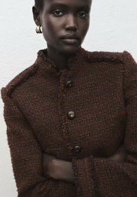 Bruine geweven jas met een gestructureerde stof, voorzien van een hoge kraag, rafelige randen en grote knopen. Lange mouwen eindigen in een uitlopende vorm.