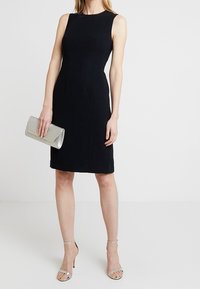 Svart ärmlös klänning i en figurnära design med vertikala sömmar, matchad med silverfärgade högklackade sandaler och en metallisk kuvertväska.