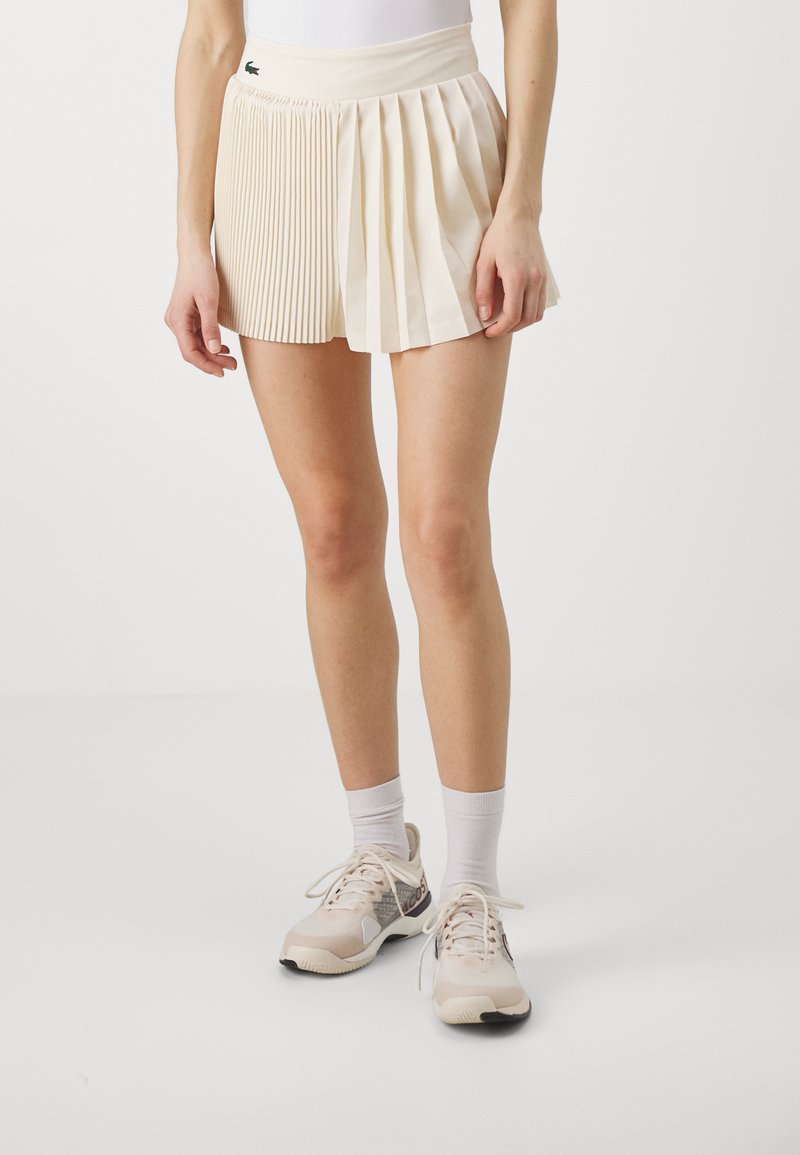 Shorts plissés crème avec une large taille, présentant des plis verticaux sur un côté et un devant lisse, agrémentés d'un logo Lacoste.