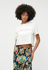 T-shirt blanc coupé en coton, avec un motif floral rose sur la poitrine ; assorti à un pantalon taille haute coloré et à motifs.