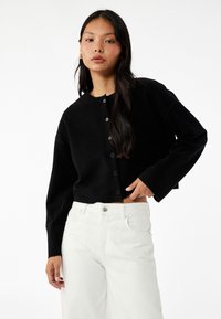 Schwarze, kurz geschnittene Strickjacke mit rundem Ausschnitt und Knöpfen vorne. Weicher Stoff, lange Ärmel, kombiniert mit weißen, hoch taillierten Hosen.