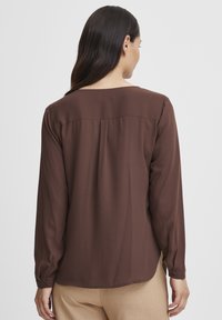 Blusa marrone a maniche lunghe con una texture liscia, scollatura rotonda e dettaglio a pieghe sul retro. Indossata con pantaloni beige.