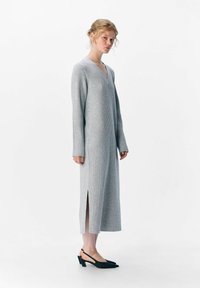 Abito a maglia grigio a coste con maniche lunghe e scollatura a V, caratterizzato da spacchi laterali e abbinato a décolleté nere con tacco.