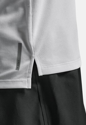 Vit sportskjorta med ett strukturerat rutmönster, sidofickor och en grå logotypdetalj. Skjortan är kombinerad med svarta shorts.