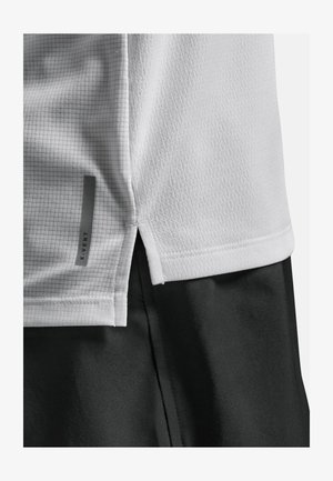 Vit sportskjorta med ett strukturerat rutmönster, sidofickor och en grå logotypdetalj. Skjortan är kombinerad med svarta shorts.