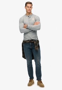 Man die met gekruiste armen staat, gekleed in een grijs langarm-shirt, blauwe jeans, tankleurige laarzen, een horloge en een geruit shirt dat rond zijn taille is geknoopt, tegen een witte achtergrond.