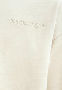 Heled beige kapuutsiga särk, millel on peen tekstuur. Omab tikitud "ORIGINALS™" logo sobivas värvitoonis õlavarre õmbluse lähedal.