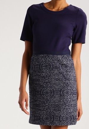 Femme portant une robe bleu marine à manches courtes avec un haut uni et une jupe à motifs complexes bleu foncé et gris.