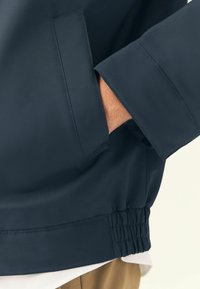 Giacca a vento navy con materiale liscio, polsini elasticizzati e tasche laterali, caratterizzata da dettagli arricciati in vita e vestibilità sartoriale.