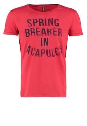 Mężczyzna w czerwonej koszulce z napisem "Spring Breaker in Acapulco", niebieskich jeansach, szarych trampkach, trzymający szary sweter, stojący z lekko uniesioną jedną nogą.
