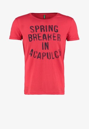 Man draagt een rood T-shirt met de tekst "Spring Breaker in Acapulco", blauwe spijkerbroek, grijze sneakers, houdt een grijze trui vast en staat met één been iets opgetild.