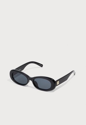 Lunettes de soleil noires en forme ovale avec des verres sombres, une monture en plastique brillant, des branches fines et un petit accent doré sur le côté.