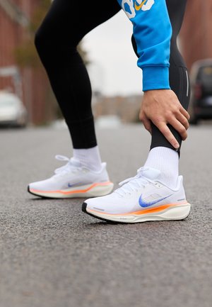 Biegacz w białych i pomarańczowych butach do biegania Nike oraz czarnych legginsach rozciąga nogę na ulicy miejskiej.
