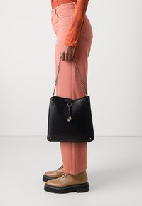 Czarna skórzana torba typu bucket z teksturowanym wykończeniem, złotymi elementami i łańcuszkowym paskiem. Noszona z jasnoróżowymi spodniami i beżowymi butami.