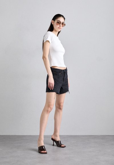 Žena oblečená v bielom priliehavom tričku s krátkym rukávom, čiernych džínsových šortkách, hnedých sandáloch na podpätku a s okuliarmi stojí na svetlosivom pozadí.