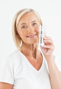 Silvrig cylindrisk hudvårdsserumbekar som hålls av en kvinna, med tydlig märkning och en glänsande yta, mot en vit bakgrund.