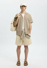 Beige textuur korte mouwen shirt, witte t-shirt, beige shorts, grijze trui gedrapeerd, en olijfgroene sandalen. Een bijpassende beige pet en tas.