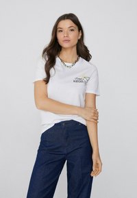 Weißes T-Shirt mit gesticktem Design "Ich brauche einen Margarita", getragen mit dunkelblauen Jeans. Einfacher, lässiger Schnitt.