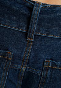 Mörkblå denimjeans med orange sömnad, utrustad med två bakfickor och en synlig midjebandsslinga för extra struktur.