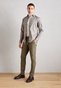 Camisa de botones con ligero estampado, cuello alto beige, pantalones verde oliva y mocasines de cuero negro. Fondo de suelo de madera natural.