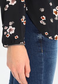 Blouse noire à motifs floraux avec de longues manches froncées, ornée de fleurs roses et marron. Associée à un jean en denim bleu, offrant une coupe ajustée.