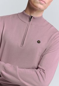 Lichtroze half-zip pullover van zachte brei-stof met lange mouwen. Heeft een hexagonaal logo-opplanation op de borst en een hoge kraag.