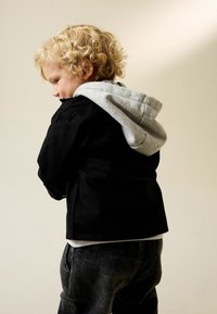 Črna denim jakna z sivo kapuco, oblečena čez belo srajco. Kodraste blond lase otroka je mogoče videti od zadaj. Temno sive kavbojke zaključujejo oblačilo.