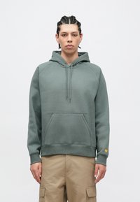 Zeleni hoodie s vezicama, kangaroo džepom i rebrastim manžetnama. Tkanina izgleda mekano i teksturirano, s naglaskom na logotip na rukavu.