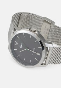 Reloj con correa de malla de plata y un dial gris oscuro, que presenta marcadores de hora blancos, una aguja de segundos verde y un logo de Lacoste en el centro.