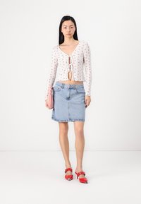 Hvit langermet topp med blomstertrykk, kort design og knytfront, kombinert med et lys blått denimm skjørt og røde spisse hæler.
