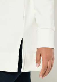 Sudadera de algodón blanca con aberturas laterales, puños acanalados y una textura suave. El dobladillo es ligeramente más largo en la parte trasera que en la delantera.