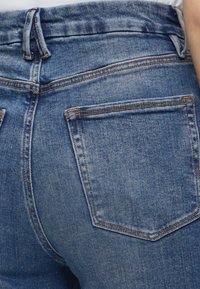 Κοντινό στην πίσω τσέπη μπλε τζιν με πορτοκαλί ραφές και θηλιές ζώνης σε άτομο που φοράει λευκό τοπ.