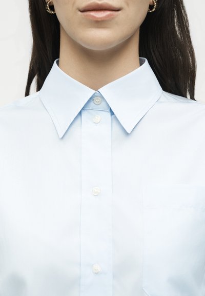 Γυναίκα με ανοιχτό μπλε πουκάμισο με κουμπιά και μυτερό γιακά, και χρυσούς κρίκους στα αυτιά, εμφανιζόμενη από το πηγούνι μέχρι το στήθος.