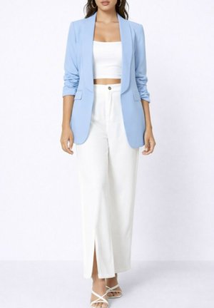 Femme portant un blazer bleu clair, un haut court blanc, un pantalon blanc taille haute à jambes larges avec une fente à l'avant, et des talons blancs à lanières.