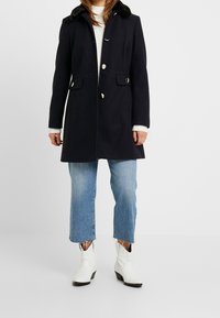 Cappotto in misto lana blu navy con colletto in pelliccia sintetica, dotato di bottoni argentati, due tasche frontali e una vestibilità sagomata sopra jeans a 3/4 di un blu chiaro.