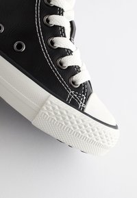 Baskets montantes noires avec semelle en caoutchouc blanche, bout en caoutchouc texturé, lacets blancs et coutures contrastantes le long des côtés.