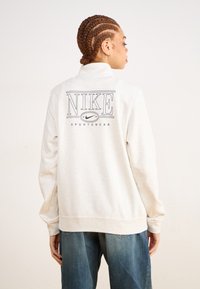 Lys grå Nike hettegenser med høy krage, med en bold logo og "SPORTSWEAR" tekst på baksiden. Mykt stoff, avslappet passform.